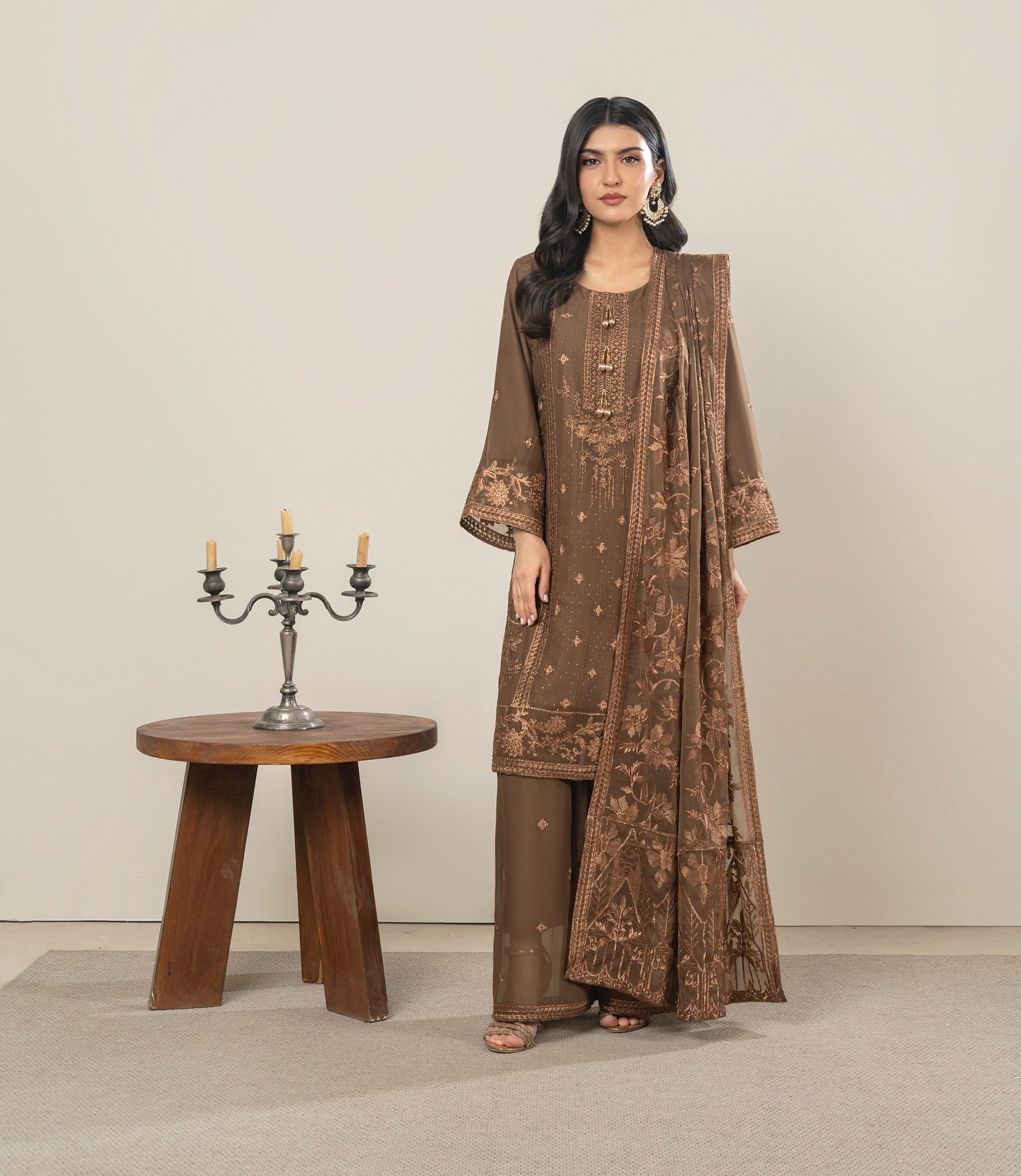 Woman in a brown embroidered outfit standing next to a wooden table with a candle holder.