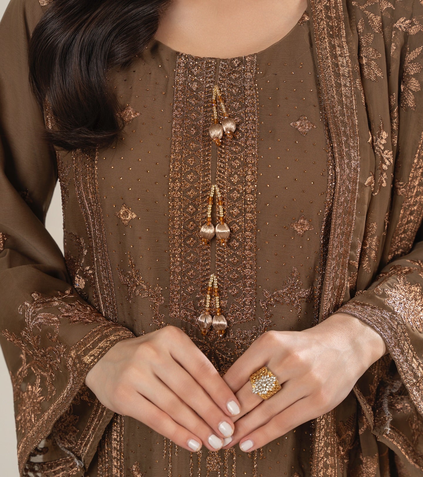 Close-up of a person wearing a brown embroidered outfit with a ring on a neutral background