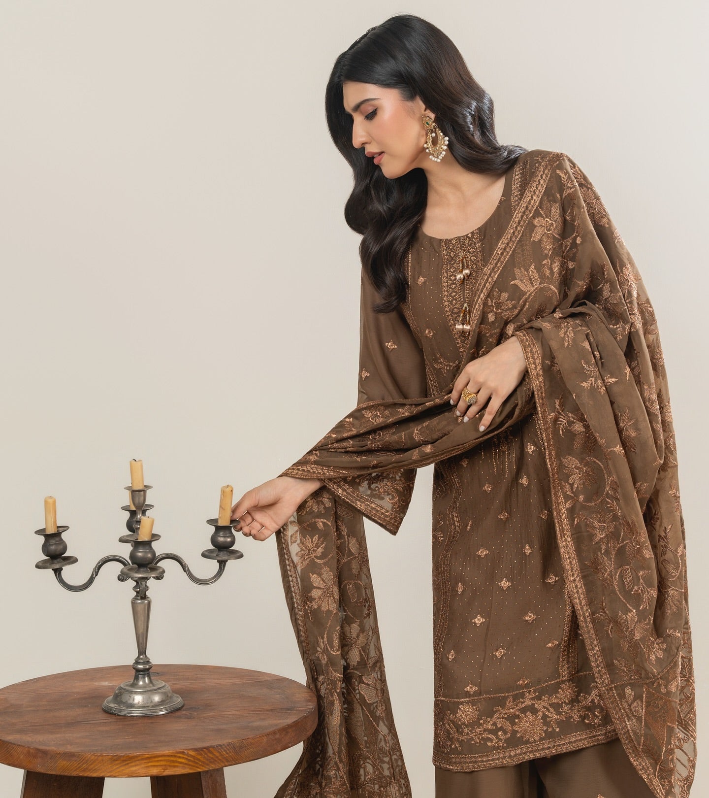 Woman in a brown embroidered outfit standing next to a wooden table with a candle holder.