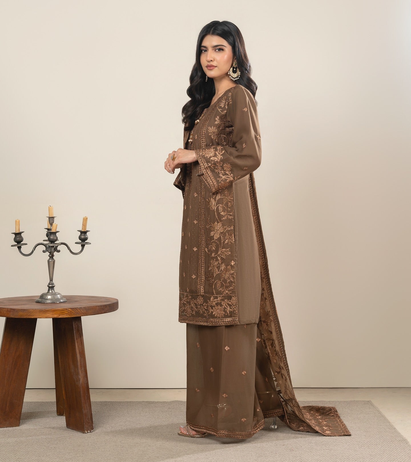 Woman wearing a brown embroidered outfit standing next to a wooden table with candles.